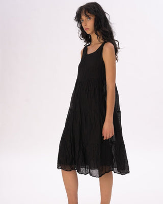 A woman standing against a plain background, wearing a black knee-length dress with a tiered waist and crinkled texture, paired with a black top and flat shoes.