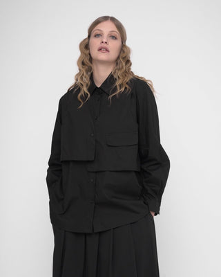 A woman wearing a black buttoned long sleeve shirt with a layered crop shirt over the top, paired with a dark skirt.