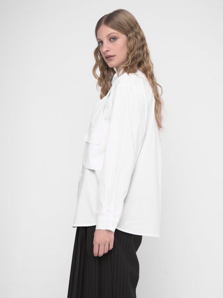 A woman wearing a white buttoned long sleeve shirt with a layered crop shirt over the top, paired with a dark skirt.