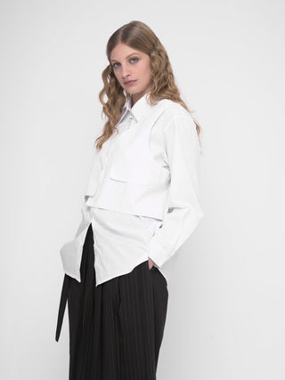A woman wearing a white buttoned long sleeve shirt with a layered crop shirt over the top, paired with a dark skirt.