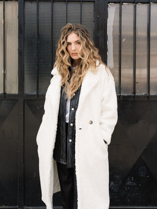 A woman standing in front of a black metal gate, wearing a white double-breasted overcoat with a flat collar, button closures, and slant pockets. The coat reaches below the knee.
